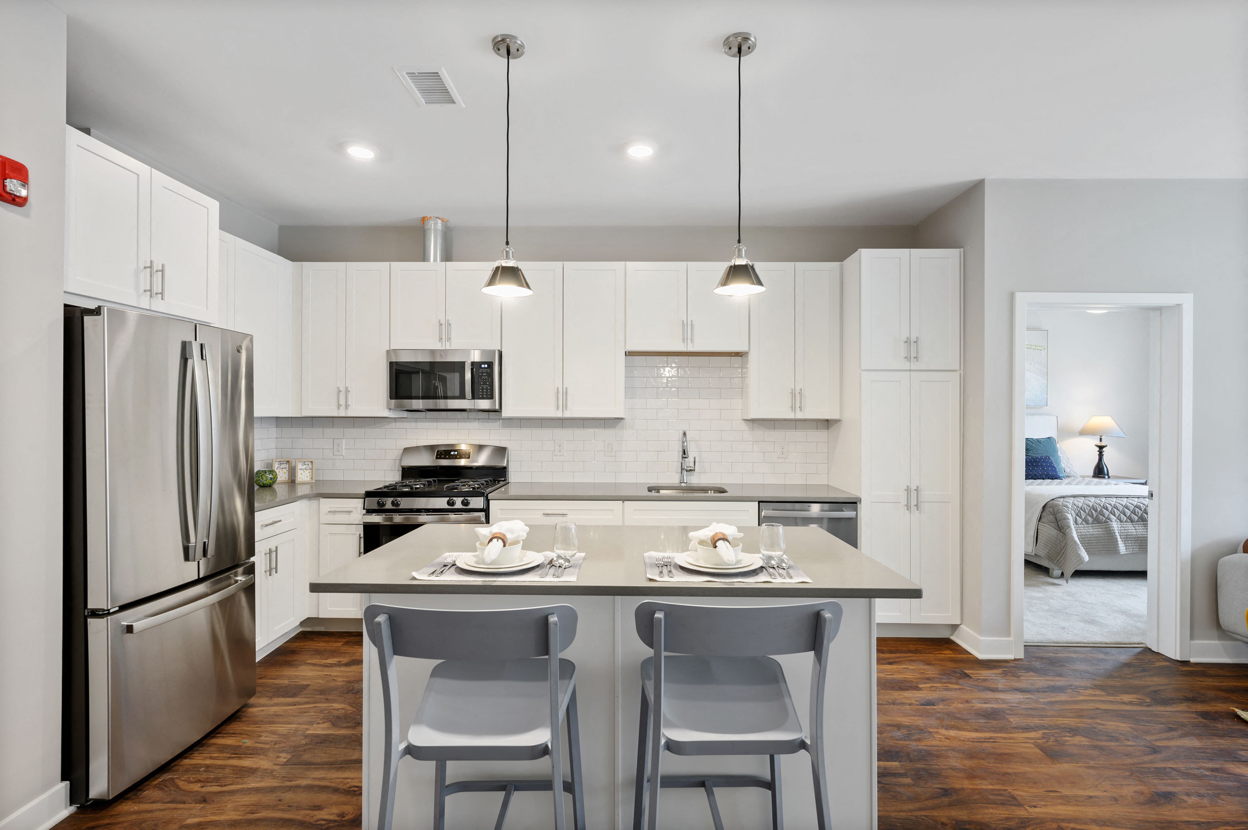 A large L-shaped kitchen with pendant and recessed lighting, hardwood-style flooring, stainless steel appliances, and kitchen island near a bedroom.