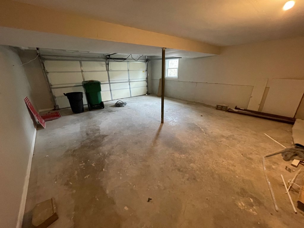 A room with a concrete floor and a window with a view of a green trash can and a red ladder.