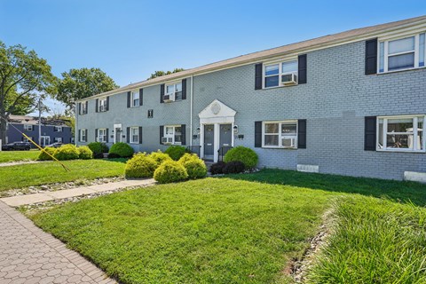 A building with a white door and black shutters is surrounded by green grass and bushes.