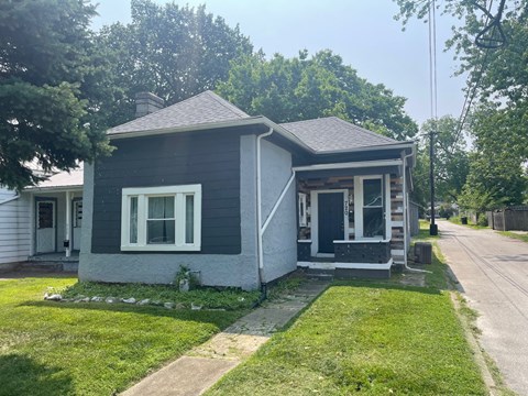 A small house with a grey roof and white trim is surrounded by a grassy lawn.