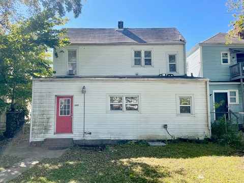 A white house with a red door is in front of a tree.