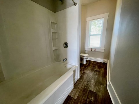 A white bathtub with a shower head and a toilet in a bathroom with wood flooring.