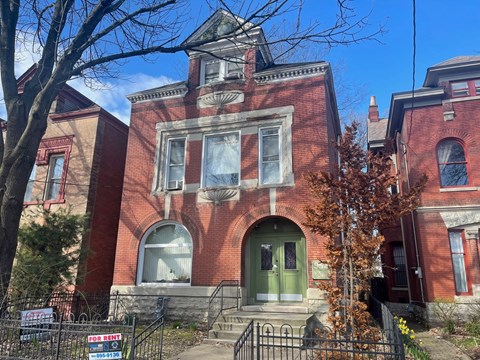 A red brick building with a green door and a "For Rent" sign in front.