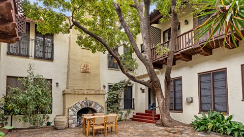 A patio with a table and chairs is surrounded by a tree and a building.