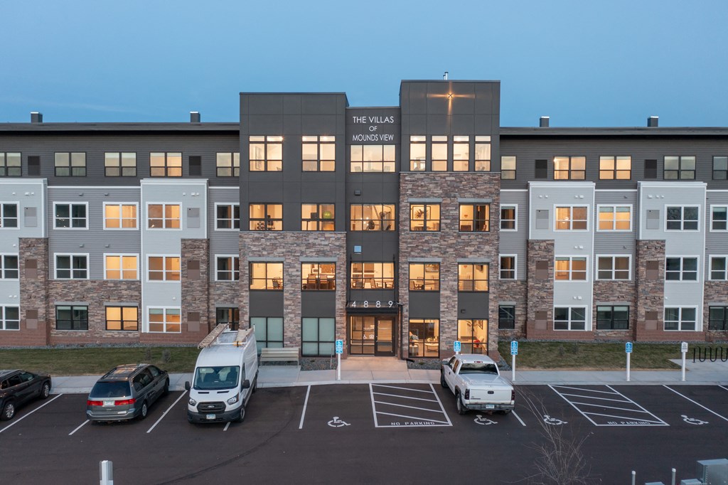 an exterior view of an apartment building at dusk with cars parked in front