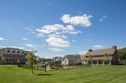 A playground with a slide is in the foreground of a grassy field with houses in the background.