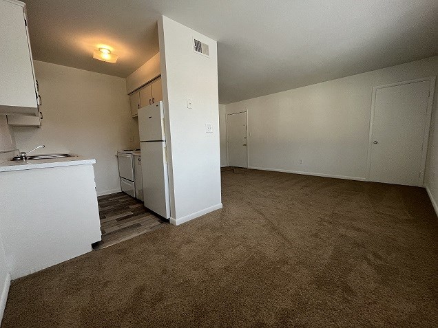 an empty living room with a kitchen and a refrigerator