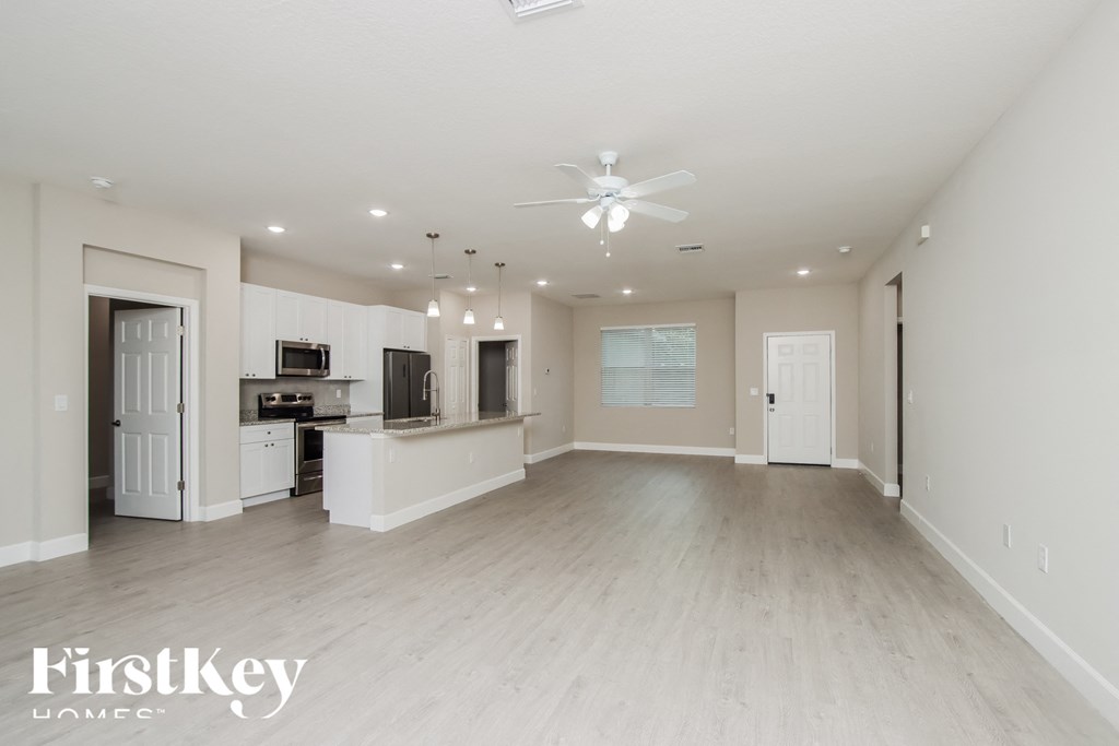 A spacious living room with a kitchen in the background and a ceiling fan in the middle of the room.
