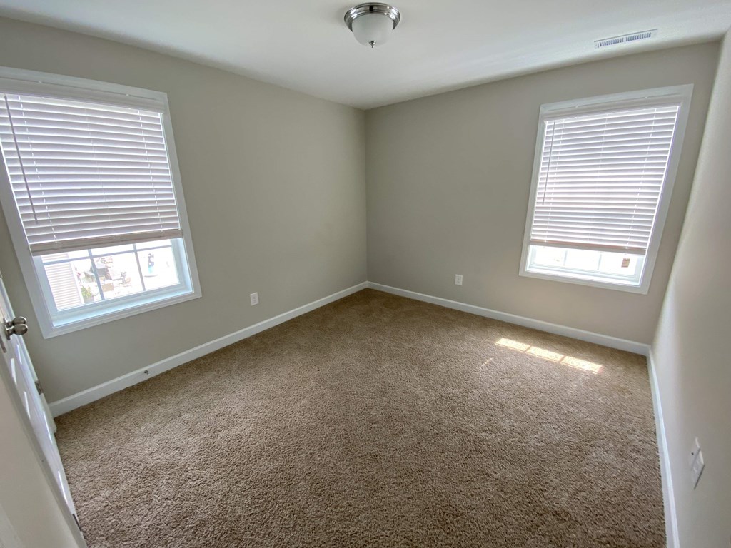 A room with carpeted floor, two windows with blinds, and a ceiling fan.