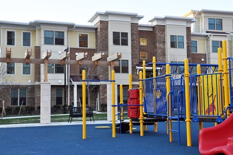 A playground with a blue surface and yellow and red play equipment in front of apartment buildings.