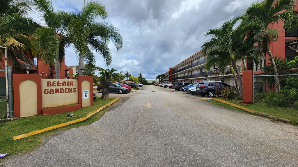 A road leading to a red building with the sign "Belair Gardens" on it.
