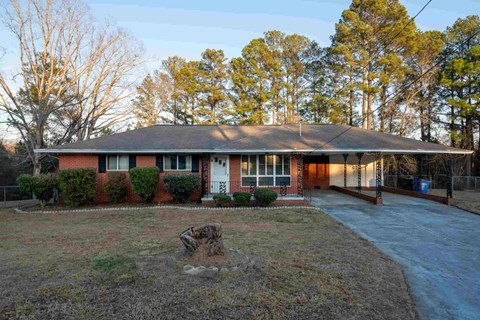 A house with a red brick exterior and a grey roof is surrounded by a grassy area and trees.