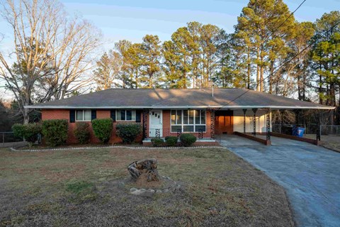 A house with a red brick exterior and a grey roof is surrounded by a grassy area and trees.