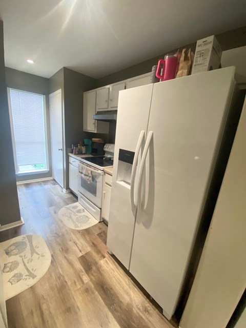 A white refrigerator stands in a kitchen with a wooden floor and a window.