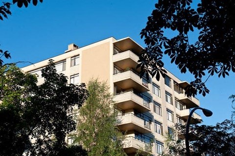 A tall beige building with balconies is surrounded by trees.