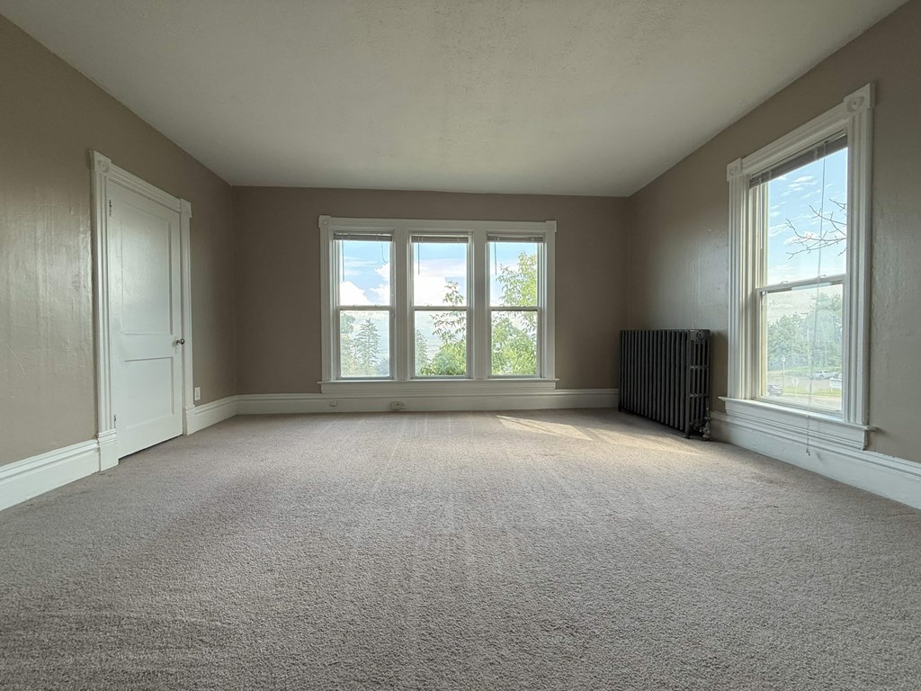 A room with a carpeted floor, a window with a view of trees, and a radiator.