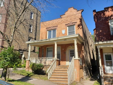 A red brick house with a porch and a tree in front.