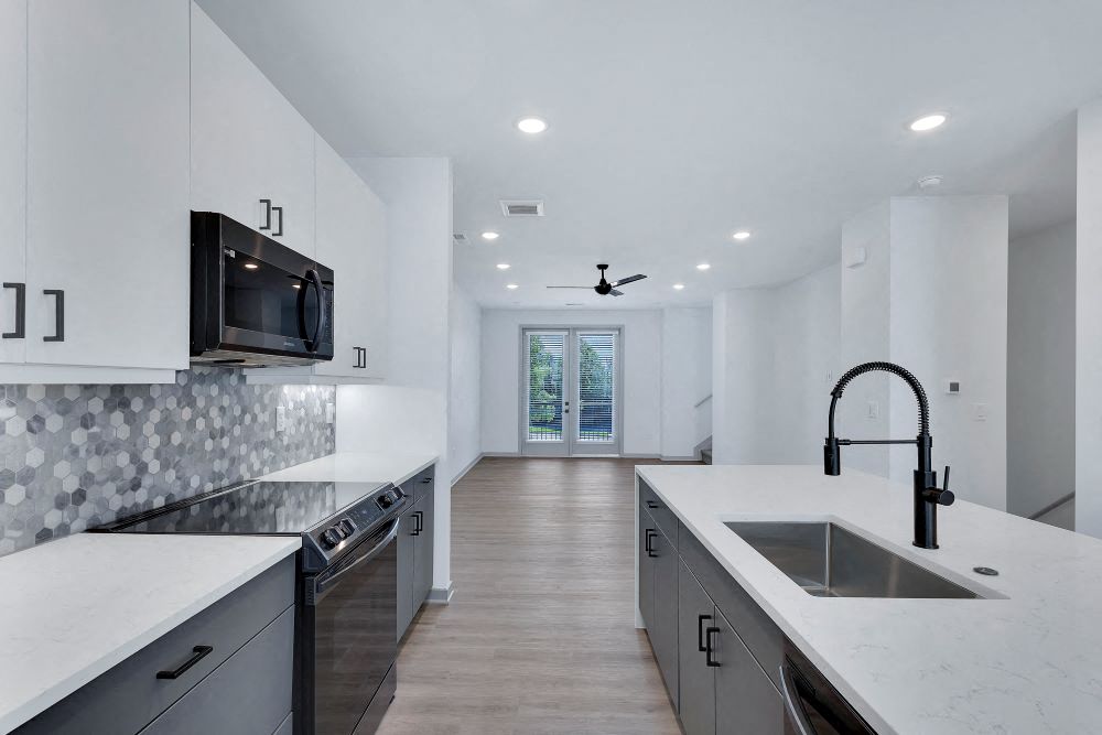 Close up view of kitchen with large waterfall island and sink, tile backsplash, soft closing cabinets and black stainless steel appliances