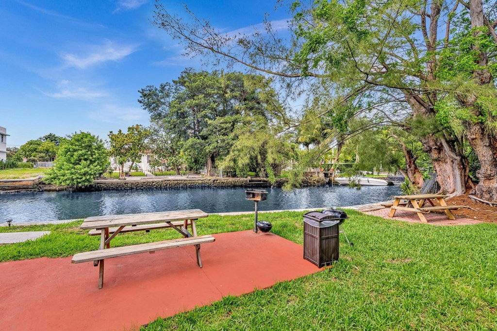 A picnic table sits on a red pathway next to a trash can in a park.