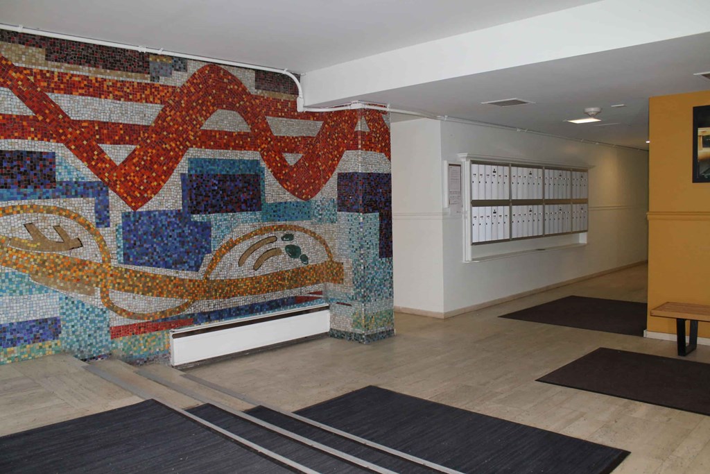 A large mosaic wall in a hallway with a bench and rugs on the floor.