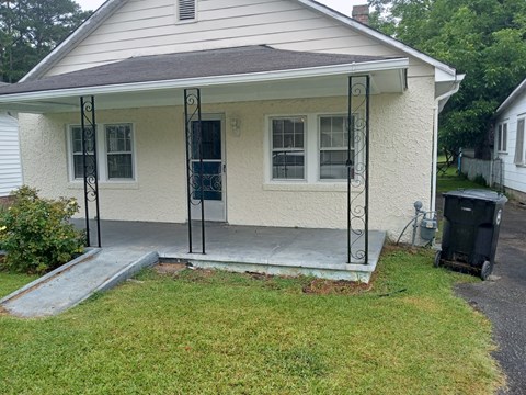 A small house with a porch and a trash can in front.