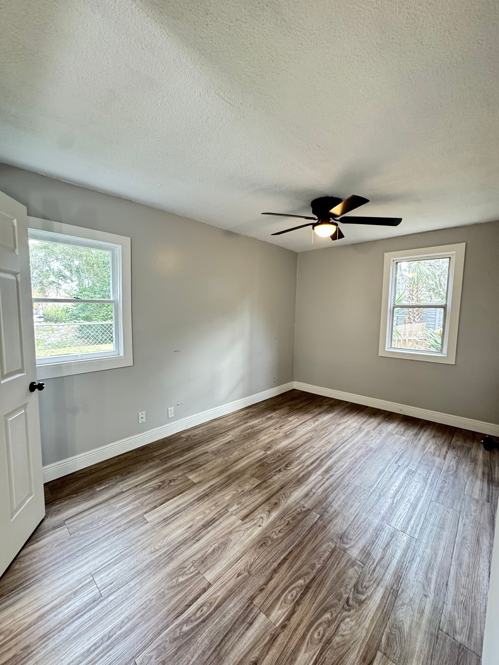 A room with a ceiling fan and light, hardwood floors, and a window with a view of a tree and a fence.