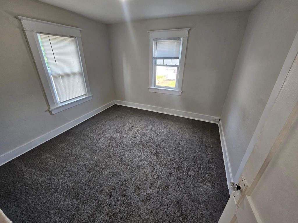 A room with grey carpet and two windows.