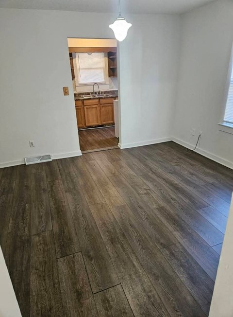 A room with wooden flooring and a kitchen visible through the door.