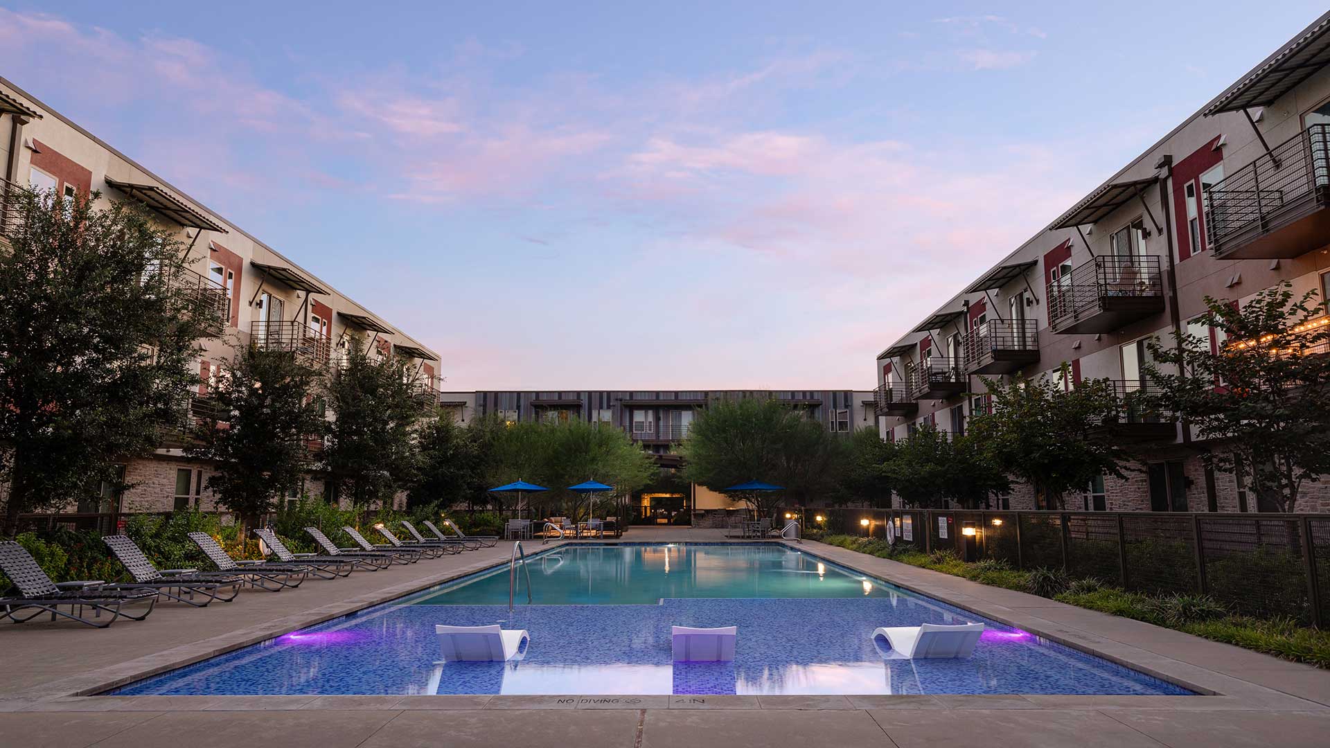 An apartment complex with a pool and lounge chairs at dusk.