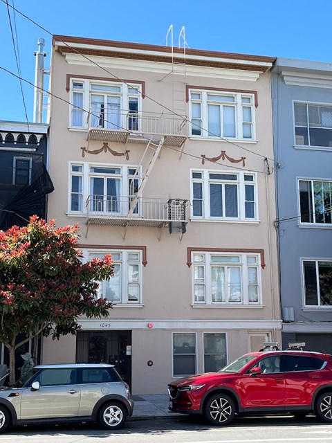A red car is parked in front of a two-story building with a balcony.