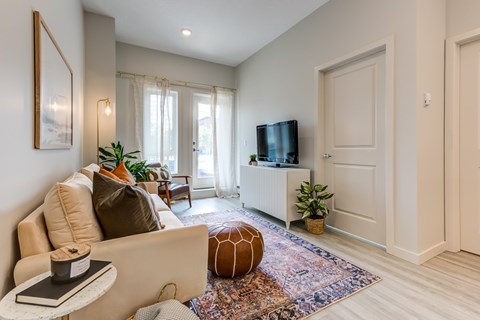 A living room with a beige sofa, a small table with a book and a jar on it, a television, a window with curtains, and a rug with a geometric design.