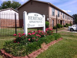 a sign for heritage apartments in front of a fence and flowers