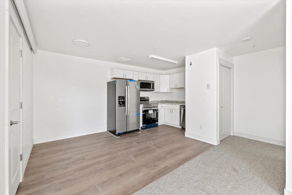 an empty living room with a kitchen with a refrigerator