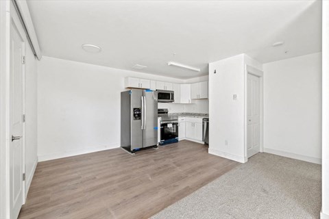 an empty living room with a kitchen with a refrigerator