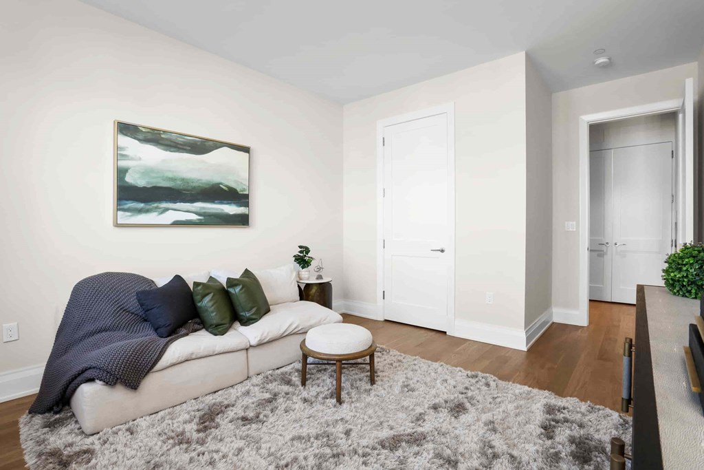 A living room with a grey rug, a grey couch with a blue blanket, a small wooden table and a painting on the wall.
