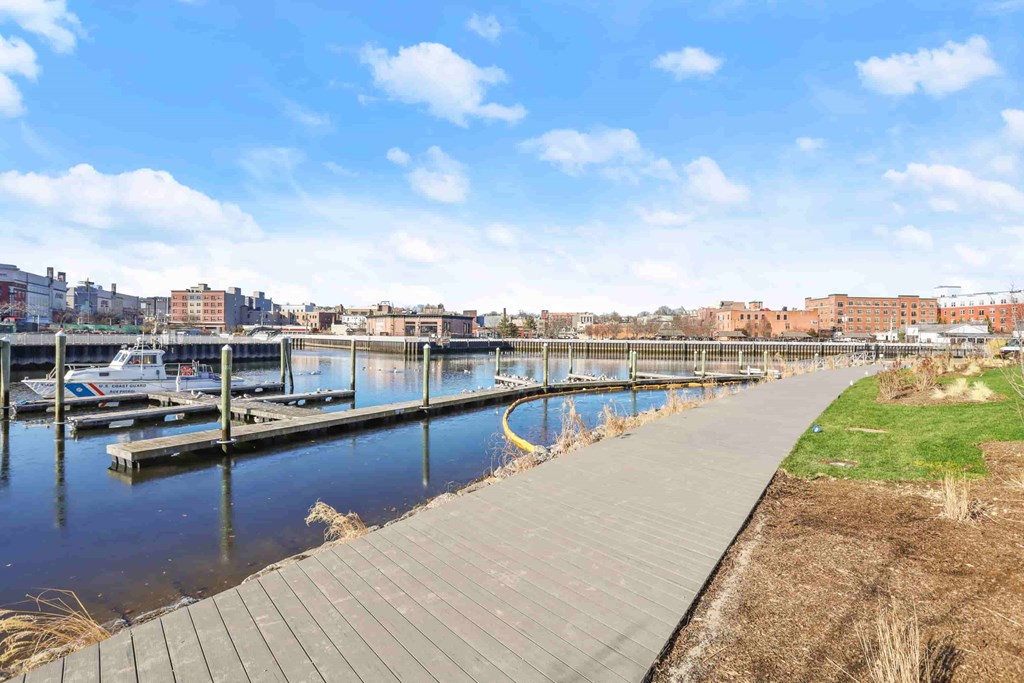 A walkway runs parallel to a body of water with buildings in the background.