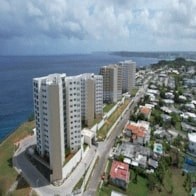 A row of white buildings are situated on a hillside overlooking the ocean.