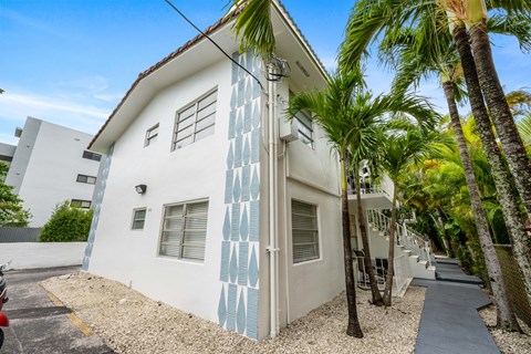 A white house with blue patterns on the side is surrounded by palm trees.