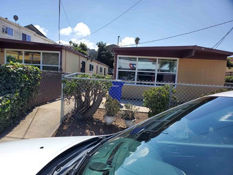 A white car is parked in front of a house with a blue sign on the fence.