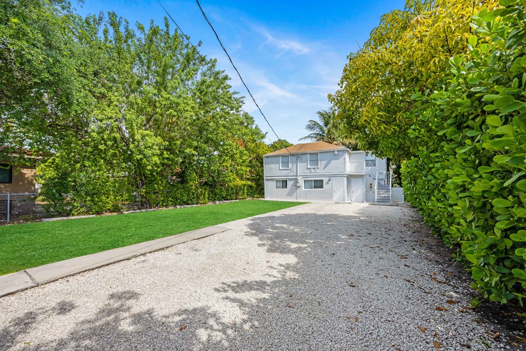 A gravel driveway leads to a white house surrounded by greenery.