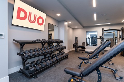 DUO Apartments gym featuring multiple adjustable benches, dumbbell racks, and a DUO-branded screen on the wall, creating a modern and organized workout space.
