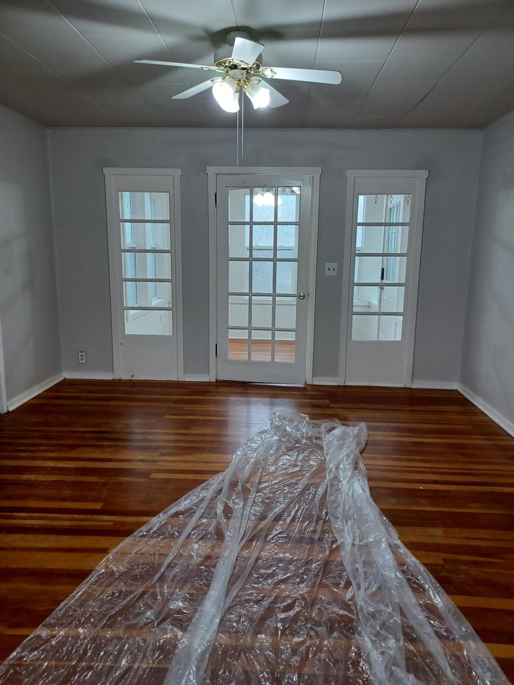 A room with a ceiling fan and wooden floors covered in plastic.