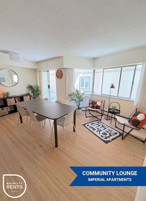 A community lounge area with a table, chairs, and a rug in a room with wooden flooring and white walls.