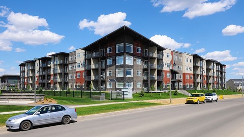 A silver car is parked on the side of a road in front of a large apartment building.