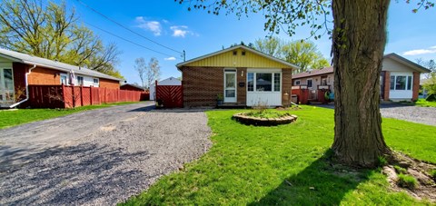 A small house with a yellow roof is surrounded by a gravel driveway and a grassy area with a tree.