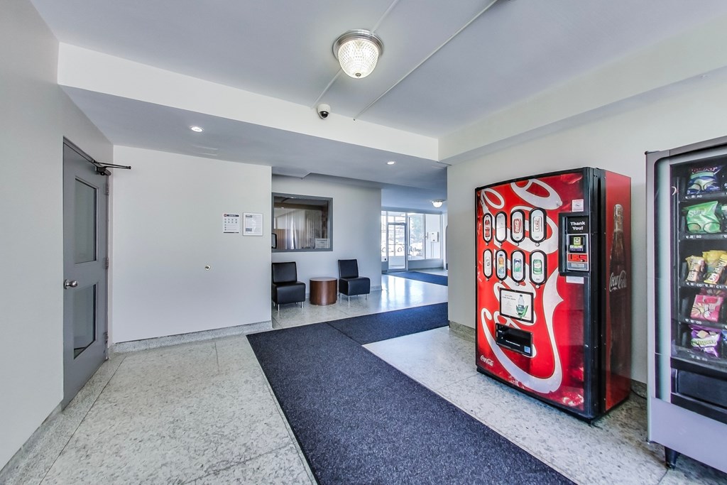 A vending machine with a red and white design is in the foreground of a room with a grey carpet.