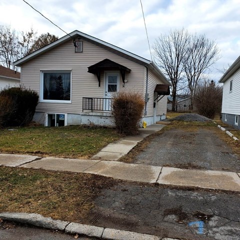 A house with a brown roof and a grey wall is surrounded by a grassy area and a paved driveway.