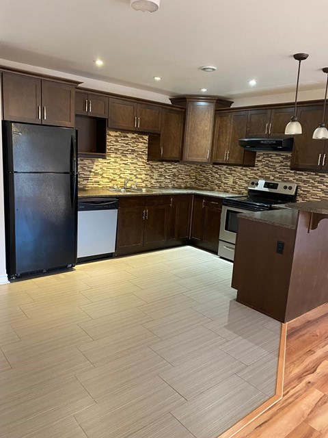 A black refrigerator stands in a kitchen with wooden cabinets and a stone backsplash.