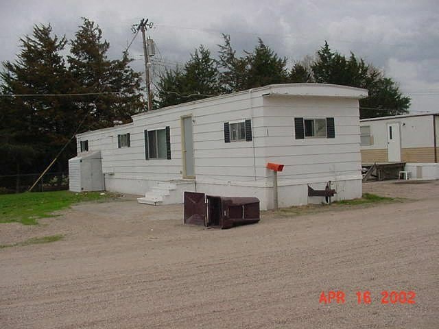 A white mobile home sits in a gravel lot with a mailbox in front of it.
