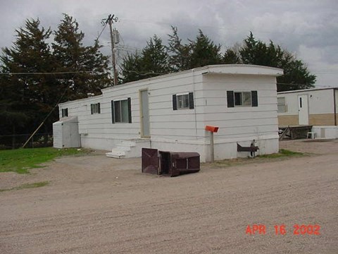 A white mobile home sits in a gravel lot with a mailbox in front of it.